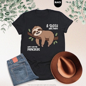 K&ouml;nnte beinhalten: Schwarzes T-Shirt mit einem Faultier-Motiv auf einem Ast und dem Text "A sloth does more work than my pancreas." Das Bild zeigt auch blaue Jeans und einen braunen Hut.