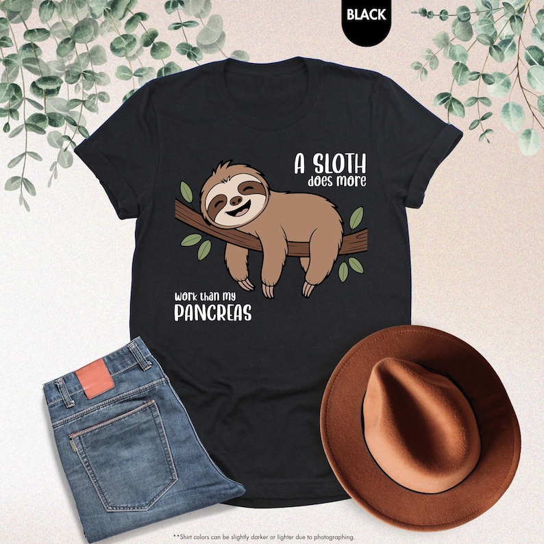 K&ouml;nnte beinhalten: Schwarzes T-Shirt mit einem Faultier-Motiv auf einem Ast und dem Text "A sloth does more work than my pancreas." Das Bild zeigt auch blaue Jeans und einen braunen Hut.