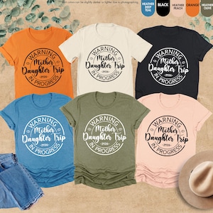 K&ouml;nnte beinhalten: Eine Auswahl an kurz&auml;rmeligen T-Shirts in verschiedenen Farben wie Orange, Creme, Schwarz, Blau, Olivgr&uuml;n und Pfirsich. Jedes Shirt zeigt einen runden Aufdruck mit dem Text "WARNING Mother Daughter Trip -2026- IN PROGRESS".