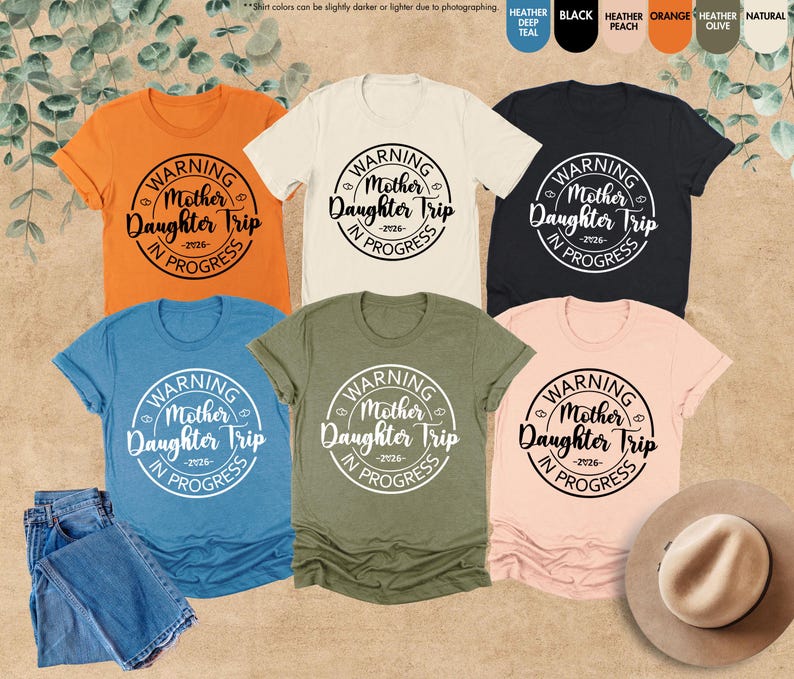 K&ouml;nnte beinhalten: Eine Auswahl an kurz&auml;rmeligen T-Shirts in verschiedenen Farben wie Orange, Creme, Schwarz, Blau, Olivgr&uuml;n und Pfirsich. Jedes Shirt zeigt einen runden Aufdruck mit dem Text "WARNING Mother Daughter Trip -2026- IN PROGRESS".