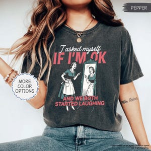 May include: Dark gray t-shirt with the text "I asked myself IF I'M OK and we both started laughing" in white and pink. The graphic shows a woman looking in a mirror. The shirt has a vintage, worn look.