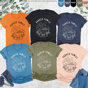 May include: Several t-shirts in shades like orange, black, and blue. Each shirt displays a white graphic with the text "PARKER FAMILY ROAD TRIP 2026" and a mountain and lake design. Some shirts are folded, and a pair of boots is visible.