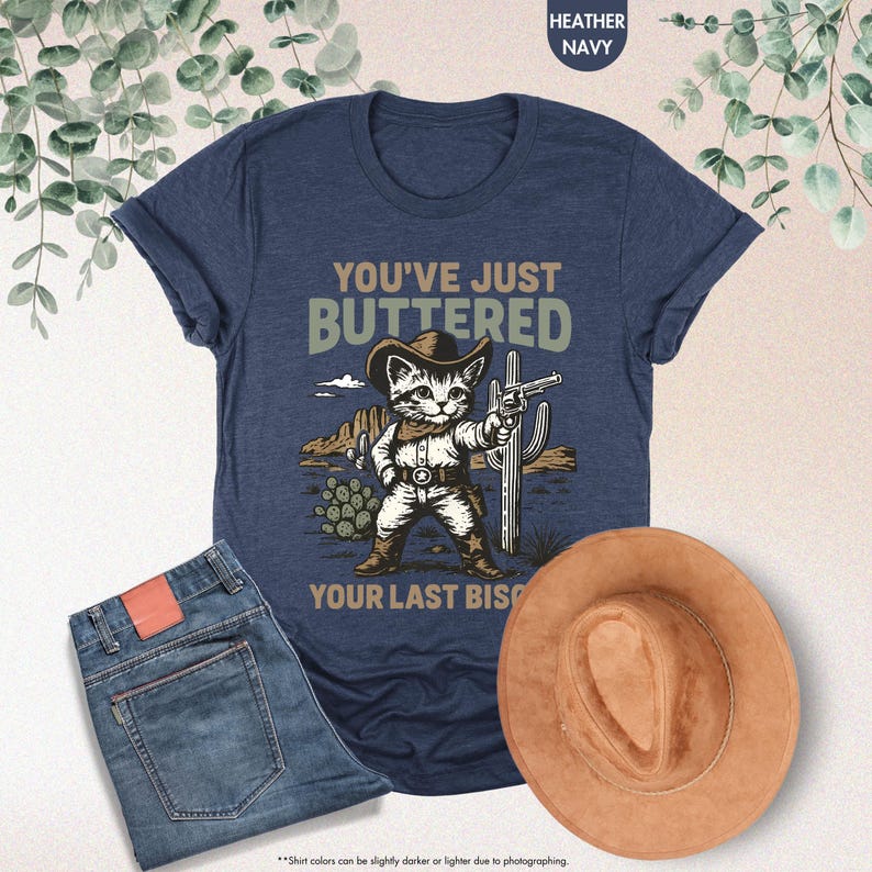 May include: Heather navy t-shirt featuring a cartoon cat in a cowboy hat holding a gun, with the text "YOU'VE JUST BUTTERED YOUR LAST BISCUIT". The image also includes blue jeans and a brown cowboy hat.