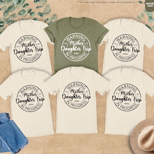 K&ouml;nnte beinhalten: Eine Auswahl an cremefarbenen und olivgr&uuml;nen T-Shirts mit dem Text "WARNING Mother Daughter Trip -2026- IN PROGRESS" in einem kreisf&ouml;rmigen Design. Eine blaue Jeans und ein Hut sind ebenfalls zu sehen.