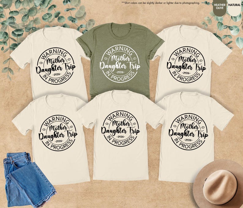 K&ouml;nnte beinhalten: Eine Auswahl an cremefarbenen und olivgr&uuml;nen T-Shirts mit dem Text "WARNING Mother Daughter Trip -2026- IN PROGRESS" in einem kreisf&ouml;rmigen Design. Eine blaue Jeans und ein Hut sind ebenfalls zu sehen.