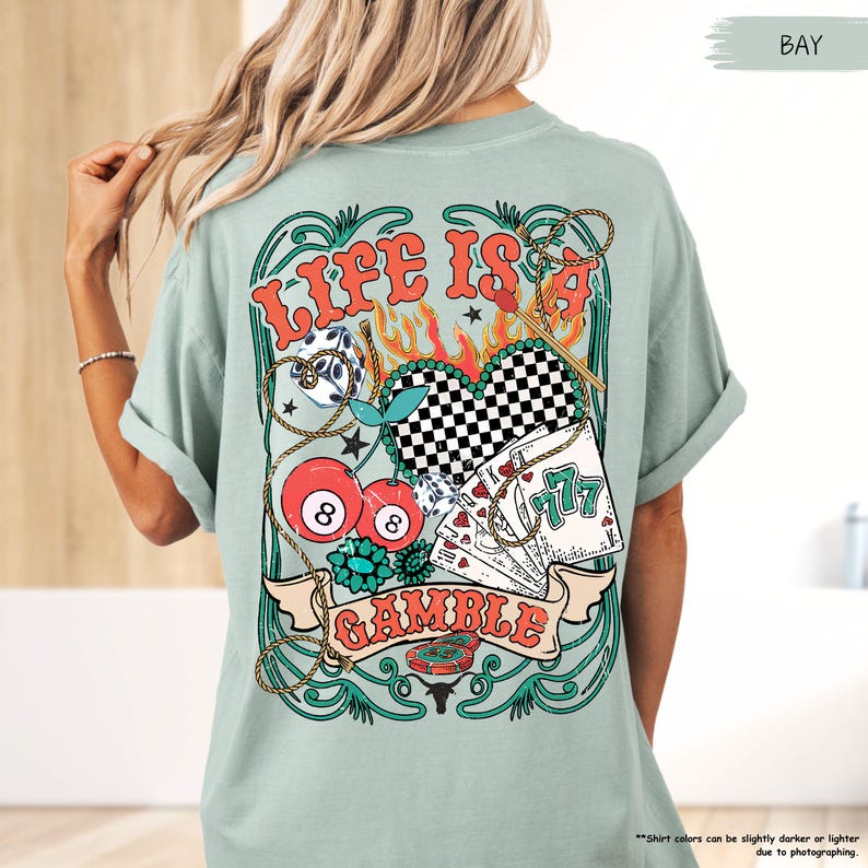 Puede incluir: Una camiseta azul claro con un dise&ntilde;o gr&aacute;fico de un coraz&oacute;n, cerezas, dados, cartas de juego y una pancarta que dice "Life is a Gamble".