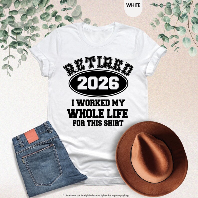 Puede incluir: Camiseta blanca con el texto negro "RETIRED 2026 I WORKED MY WHOLE LIFE FOR THIS SHIRT". Se combina con vaqueros azules y un sombrero marr&oacute;n. El texto est&aacute; en negrita, estilo universitario.