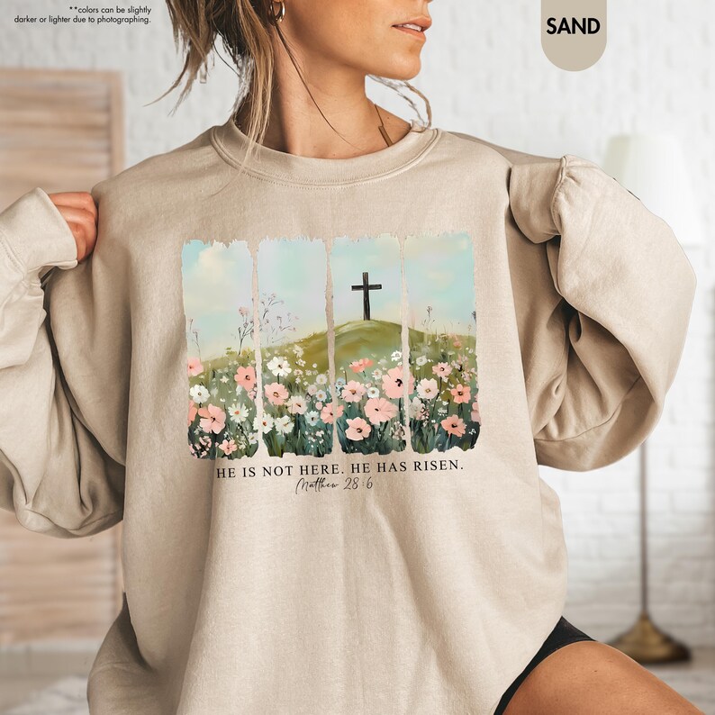 May include: A beige sweatshirt with a floral design and a cross on a hill. The text "HE IS NOT HERE. HE HAS RISEN. Matthew 28:6" is printed below the design.