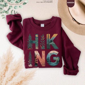 Pode incluir: Camisola de cor vinho com um design gr&aacute;fico da palavra "HIKING" com cenas da natureza incorporadas nas letras.