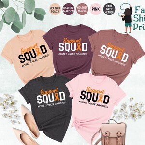 May include: Five t-shirts in different colors with the text "Support Squad" and a ribbon design. The text "#Kidney Cancer Awareness" is below the design. The t-shirts are available in heather peach, heather maroon, heather mauve, pink, and dark grey heather.