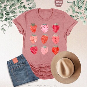 May include: A heather mauve t-shirt with a graphic of nine pink and red strawberries. The shirt is displayed on a flat surface with a pair of blue jeans and a brown hat.