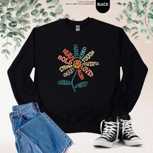 Pode incluir: Camisola preta com um design floral composto por palavras como "corajoso", "ousado" e "forte". O design &eacute; em v&aacute;rias cores, incluindo turquesa, laranja e amarelo. A camisola &eacute; combinada com jeans azuis e t&ecirc;nis pretos e brancos.