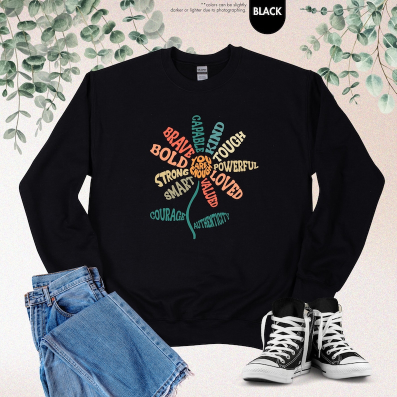 Pode incluir: Camisola preta com um design floral composto por palavras como "corajoso", "ousado" e "forte". O design &eacute; em v&aacute;rias cores, incluindo turquesa, laranja e amarelo. A camisola &eacute; combinada com jeans azuis e t&ecirc;nis pretos e brancos.