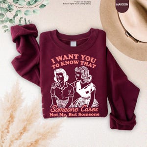 May include: Maroon sweatshirt with the text "I WANT YOU TO KNOW THAT Someone Cares Not Me, But Someone" and a graphic of two women. The sleeves are cuffed. The word "MAROON" is in the upper right corner.