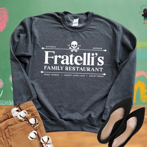 May include: A dark grey long-sleeved sweatshirt with a white graphic design that says "Fratelli's Family Restaurant" with a skull and crossbones logo. The text "Astoria, Oregon" is above the logo and the text "Dead Things, Daddy Long Legs, Rocky Road" is below the logo.