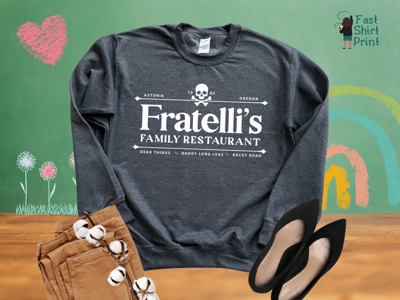May include: A dark grey long-sleeved sweatshirt with a white graphic design that says "Fratelli's Family Restaurant" with a skull and crossbones logo. The text "Astoria, Oregon" is above the logo and the text "Dead Things, Daddy Long Legs, Rocky Road" is below the logo.
