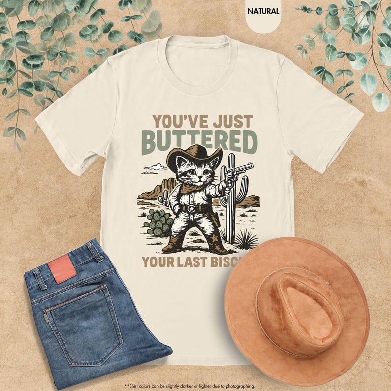 May include: Cream-coloured t-shirt with a cowboy cat graphic, jeans, and a brown hat. The graphic features a cat in a cowboy hat holding a gun, with the text "YOU'VE JUST BUTTERED YOUR LAST BISC".