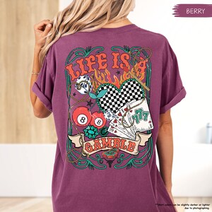 Puede incluir: Una camiseta morada con un dise&ntilde;o gr&aacute;fico de un coraz&oacute;n, dados, cartas de juego y cerezas. El dise&ntilde;o gr&aacute;fico est&aacute; rodeado por un dise&ntilde;o de cuerda verde y dorada. El texto "Life is a Gamble" est&aacute; impreso en la camiseta.