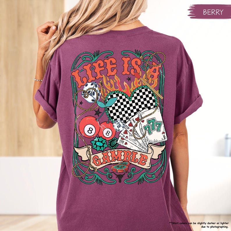 Puede incluir: Una camiseta morada con un dise&ntilde;o gr&aacute;fico de un coraz&oacute;n, dados, cartas de juego y cerezas. El dise&ntilde;o gr&aacute;fico est&aacute; rodeado por un dise&ntilde;o de cuerda verde y dorada. El texto "Life is a Gamble" est&aacute; impreso en la camiseta.