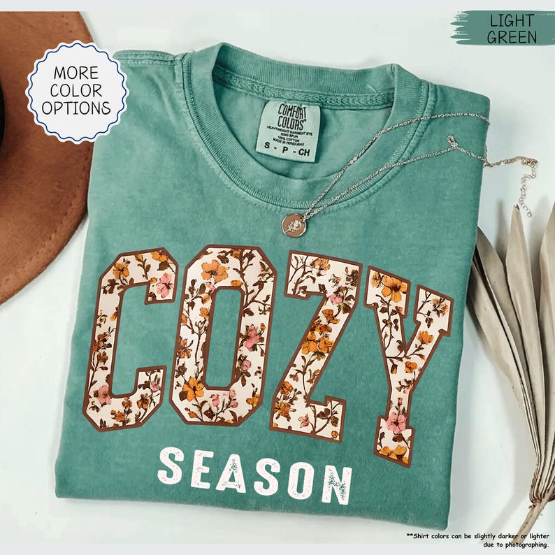 May include: A light green Comfort Colors t-shirt with the word "COZY" in large, floral-patterned letters and the word "SEASON" below. A silver necklace with a pendant rests on the shirt. The shirt is folded.