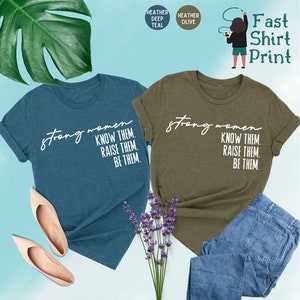 K&ouml;nnte beinhalten: Zwei T-Shirts mit dem Text "strong women know them, raise them, be them." in wei&szlig; auf einem teal- und olivgr&uuml;nen Hintergrund.