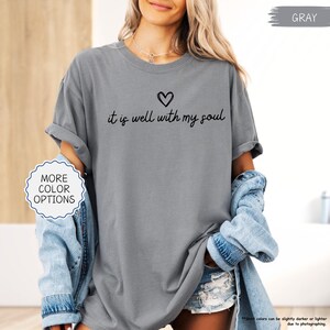 K&ouml;nnte beinhalten: Graues T-Shirt mit dem schwarzen Schriftzug "it is well with my soul" und einem Herzsymbol. Das Shirt hat hochgekrempelte &Auml;rmel. Das Bild enth&auml;lt auch einen Button "Weitere Farboptionen". Teil einer Bekleidungskollektion.