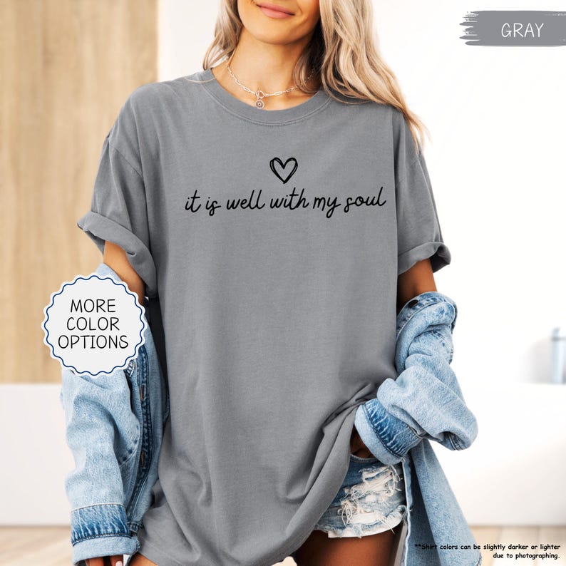K&ouml;nnte beinhalten: Graues T-Shirt mit dem schwarzen Schriftzug "it is well with my soul" und einem Herzsymbol. Das Shirt hat hochgekrempelte &Auml;rmel. Das Bild enth&auml;lt auch einen Button "Weitere Farboptionen". Teil einer Bekleidungskollektion.