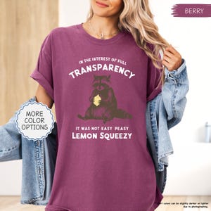 May include: Berry-colored t-shirt with the text "IN THE INTEREST OF FULL TRANSPARENCY" and "IT WAS NOT EASY PEASY LEMON SQUEEZY". The shirt features a raccoon graphic holding a lemon. The shirt has rolled-up sleeves and a crew neck.