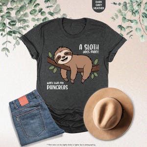 K&ouml;nnte beinhalten: Dunkelgraues Heather-T-Shirt mit einem Faultier-Motiv auf einem Ast und dem Text "A sloth does more work than my pancreas." Das Bild zeigt blaue Jeans und einen beigen Hut. Das T-Shirt ist ein Freizeitartikel.
