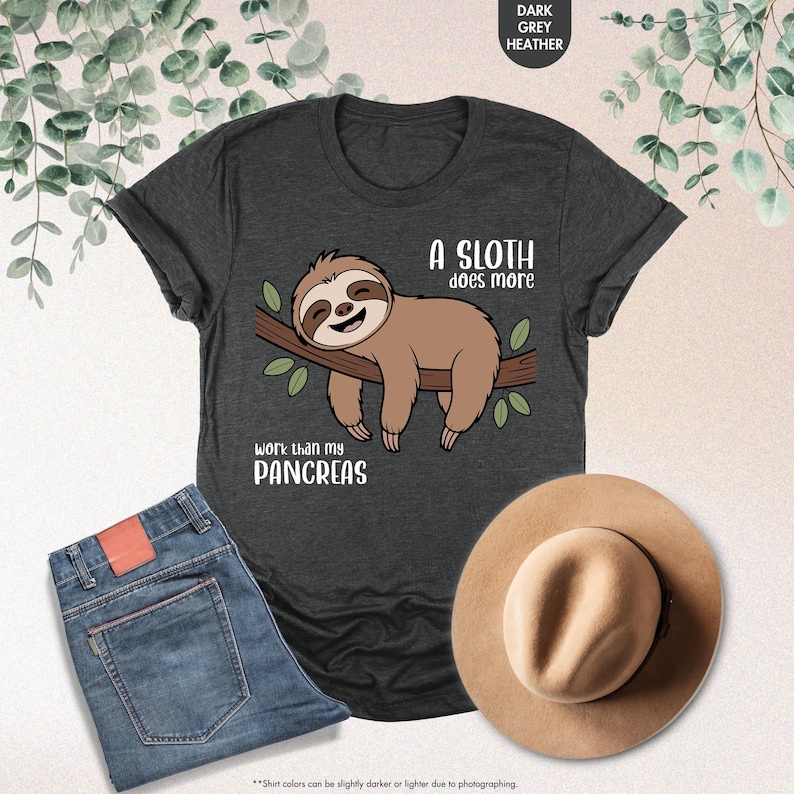 K&ouml;nnte beinhalten: Dunkelgraues Heather-T-Shirt mit einem Faultier-Motiv auf einem Ast und dem Text "A sloth does more work than my pancreas." Das Bild zeigt blaue Jeans und einen beigen Hut. Das T-Shirt ist ein Freizeitartikel.