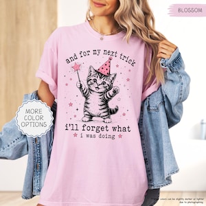 May include: A light pink t-shirt featuring a black and white illustration of a kitten wearing a pink party hat and holding a star-tipped wand. The text reads, and for my next trick i'll forget what i was doing, surrounded by small pink stars.