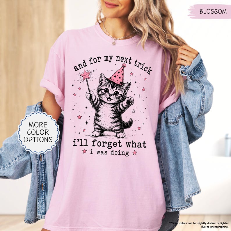 May include: A light pink t-shirt featuring a black and white illustration of a kitten wearing a pink party hat and holding a star-tipped wand. The text reads, and for my next trick i'll forget what i was doing, surrounded by small pink stars.