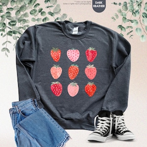 May include: A dark heather grey sweatshirt with a pattern of red and pink strawberries. The sweatshirt is shown on a model wearing blue jeans and black sneakers.