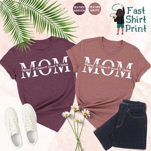 Puede incluir: Dos camisetas con la palabra "MOM" impresa en blanco sobre un fondo de color heather maroon y heather mauve. Las camisetas est&aacute;n personalizadas con nombres y corazones. El texto dice "Michael Custom Sarah Custom".