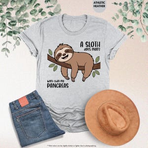 K&ouml;nnte beinhalten: Hellgraues T-Shirt mit einem gezeichneten Faultier auf einem Ast und dem Text "A sloth does more work than my pancreas". Blaue Jeans und ein brauner Hut sind ebenfalls im Bild zu sehen.