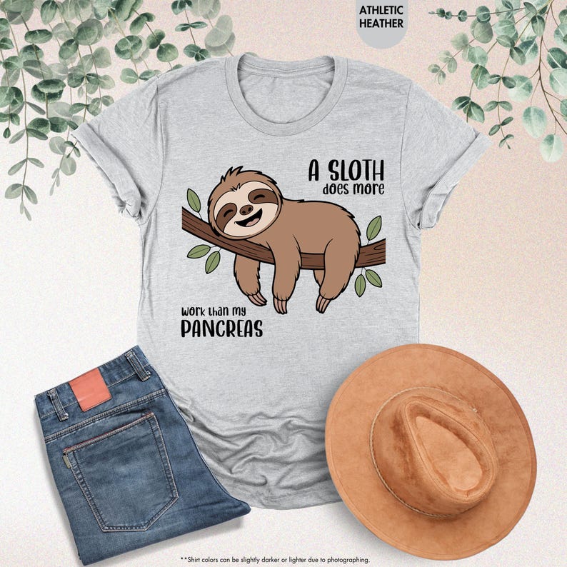 K&ouml;nnte beinhalten: Hellgraues T-Shirt mit einem gezeichneten Faultier auf einem Ast und dem Text "A sloth does more work than my pancreas". Blaue Jeans und ein brauner Hut sind ebenfalls im Bild zu sehen.