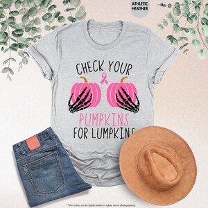 以下が含まれることがあります： 「CHECK YOUR PUMPKINS FOR LUMPKINS」の文字が黒とピンクでプリントされたライトグレーのTシャツ。デザインには、黒い骸骨の手とピンクのリボンが付いた2つのピンク色のカボチャが含まれています。青いジーンズと茶色の帽子も表示されています。
