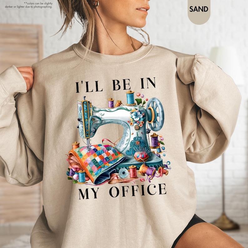 Pode incluir: Uma camisa de moletom bege com um gr&aacute;fico de m&aacute;quina de costura colorida e o texto "Vou estar no meu escrit&oacute;rio".