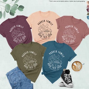 May include: Several heather t-shirts in shades like peach, mauve, maroon, olive, and deep teal. Each shirt displays a white graphic with the text "PARKER FAMILY ROAD TRIP 2026" and a mountain and lake design. The image also includes jeans and trainers.