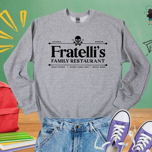 May include: A gray sweatshirt with a black graphic print of a skull and crossbones with the text "Fratelli's Family Restaurant" and the location "Astoria, Oregon". The text "Dead Things, Daddy Long Legs, Rocky Road" is printed below the restaurant name.