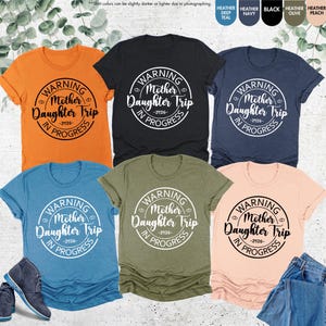 K&ouml;nnte beinhalten: Sechs T-Shirts in verschiedenen Farben, darunter Orange, Schwarz, Marine, T&uuml;rkis, Olive und Pfirsich. Jedes Shirt zeigt eine runde Grafik mit dem Text "WARNING Mother Daughter Trip -2026- IN PROGRESS" in Wei&szlig;.
