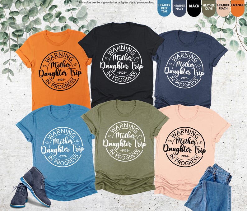 K&ouml;nnte beinhalten: Sechs T-Shirts in verschiedenen Farben, darunter Orange, Schwarz, Marine, T&uuml;rkis, Olive und Pfirsich. Jedes Shirt zeigt eine runde Grafik mit dem Text "WARNING Mother Daughter Trip -2026- IN PROGRESS" in Wei&szlig;.