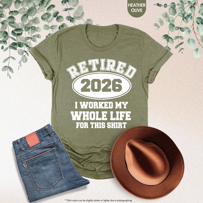 Puede incluir: Camiseta verde oliva con el texto blanco "RETIRED 2026 I WORKED MY WHOLE LIFE FOR THIS SHIRT". La imagen tambi&eacute;n incluye unos vaqueros azules y un sombrero fedora marr&oacute;n.
