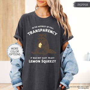 May include: Dark gray t-shirt with the text "IN THE INTEREST OF FULL TRANSPARENCY" and "IT WAS NOT EASY PEASY LEMON SQUEEZY". The graphic features a brown raccoon holding a yellow lemon. The shirt is a pepper color.