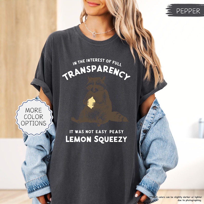 May include: Dark gray t-shirt with the text "IN THE INTEREST OF FULL TRANSPARENCY" and "IT WAS NOT EASY PEASY LEMON SQUEEZY". The graphic features a brown raccoon holding a yellow lemon. The shirt is a pepper color.