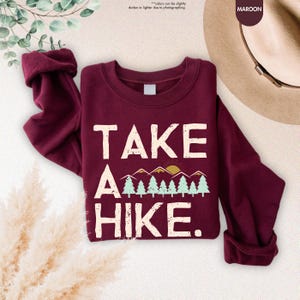 K&ouml;nnte beinhalten: Burgunderfarbenes Sweatshirt mit dem wei&szlig;en Text "TAKE A HIKE". Die Grafik zeigt Berge, B&auml;ume und eine Sonne. Das Sweatshirt hat lange &Auml;rmel und einen Rundhalsausschnitt.