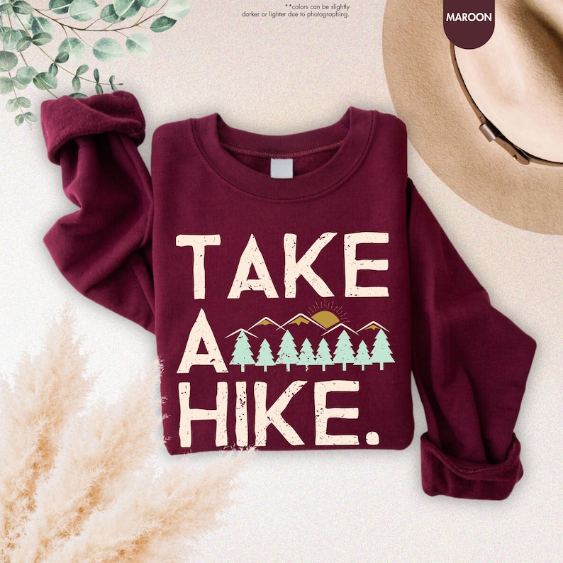 K&ouml;nnte beinhalten: Burgunderfarbenes Sweatshirt mit dem wei&szlig;en Text "TAKE A HIKE". Die Grafik zeigt Berge, B&auml;ume und eine Sonne. Das Sweatshirt hat lange &Auml;rmel und einen Rundhalsausschnitt.