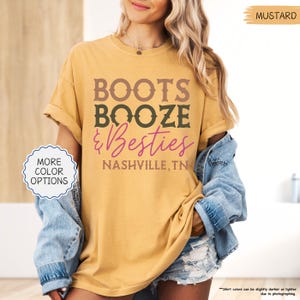 Pu&ograve; includere: T-shirt color senape con la scritta "BOOTS BOOZE & Besties NASHVILLE, TN" in lettere marroni, verdi e rosa. La maglietta ha le maniche arrotolate ed &egrave; abbinata a una giacca di jeans. Un bottone a sinistra dice "PI&Ugrave; OPZIONI DI COLORE".
