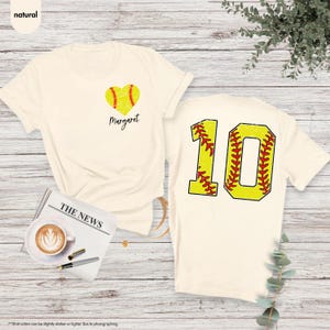 K&ouml;nnte beinhalten: Zwei cremefarbene T-Shirts mit Softball-Motiv. Ein Shirt hat ein Herz mit einem Softball darin und den Namen "Margaret" darunter. Das andere Shirt hat die Nummer "10" mit einem Softball-Design.