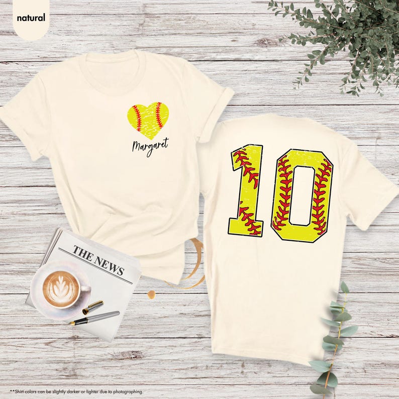 K&ouml;nnte beinhalten: Zwei cremefarbene T-Shirts mit Softball-Motiv. Ein Shirt hat ein Herz mit einem Softball darin und den Namen "Margaret" darunter. Das andere Shirt hat die Nummer "10" mit einem Softball-Design.
